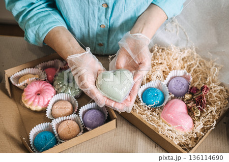 Hands hold heart shaped bath bomb over boxes filled with colorful handmade soaps and shredded paper. personalized beauty gift, diy spa kit, bespoke apothecary, aesthetic gift packaging. 136114690