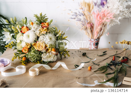 Flower bouquet featuring roses and chrysanthemums sits on brown paper near ribbon rolls. Sustainable floristry, plastic free gift wrapping, biodegradable materials, slow flower movement. 136114714