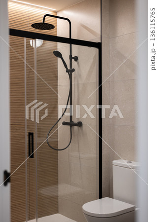 Modern bathroom with a glass shower enclosure, black fixtures, and beige tiles. A white toilet is visible in the corner. 136115765