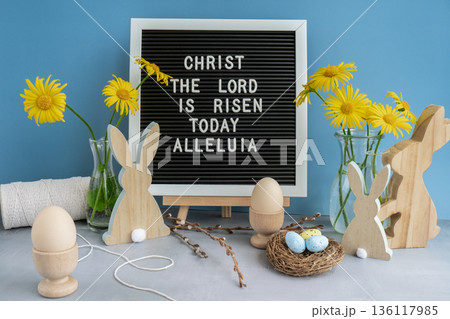 Letter board with message CHRIST THE LORD IS RISEN TODAY ALLELUIA on festive blue background and eco-friendly zero waste natural decoration. Yellow daisies flowers. Bunny egg. Holiday greeting 136117985
