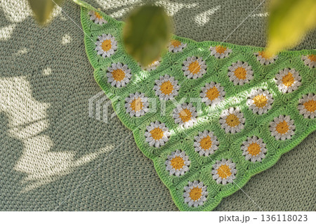 Crocheted granny square blanket featuring daisy pattern white petals and yellow center on green crochet background and houseplant leaves Crocheted granny square blanket featuring daisy pattern white petals and yellow center on green crochet background and houseplant leaves 136118023