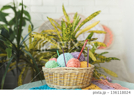 Basket filled with multi-colored balls of yarn and crochet hooks on background of home flowers Basket filled with multi-colored balls of yarn and crochet hooks on background of home flowers 136118024
