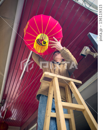 Man Hanging A Vibrant Red Lantern High Up On A Wooden Ladder Man Hanging A Vibrant Red Lantern High Up On A Wooden Ladder 136119315
