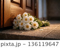 Bouquet of white daisies left on a doorstep near a wooden door in warm evening light. Concept of belated birthday greeting, apology, forgiveness and quiet regret. 136119647