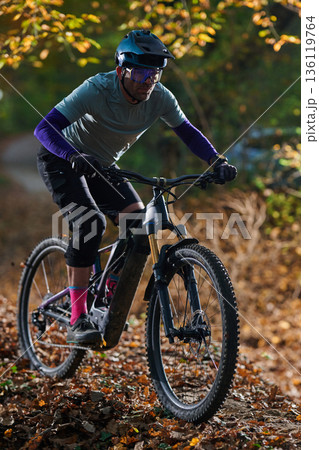 Mountain Biker Racing Down Trail in Autumn Forest With Helmet and Protective Gear During Leaf Fall Mountain Biker Racing Down Trail in Autumn Forest With Helmet and Protective Gear During Leaf Fall 136119764
