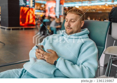Cheerful young Caucasian man sitting on a bench while holding smartphone, waiting at airport lounge lobby. 136119867