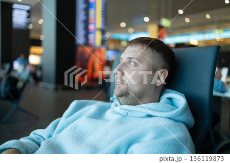 Cheerful young Caucasian man sitting on a bench while holding smartphone, waiting at airport lounge lobby. Cheerful young Caucasian man sitting on a bench while holding smartphone, waiting at airport lounge lobby. 136119873