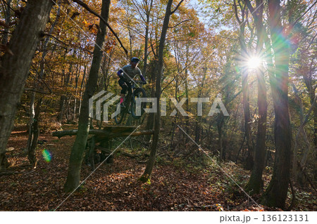 Mountain Biker Jumping Over Log in Autumn Forest Trail with Sun Burst Mountain Biker Jumping Over Log in Autumn Forest Trail with Sun Burst 136123131