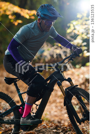 Mountain Biker Riding Through Sunlit Forest Trail in Protective Gear, Focused on Ride Mountain Biker Riding Through Sunlit Forest Trail in Protective Gear, Focused on Ride 136123535