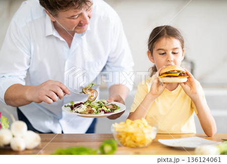 Little daughter eats a meat burger in the kitchen, her father offers her healthy vegetable salad Little daughter eats a meat burger in the kitchen, her father offers her healthy vegetable salad 136124061