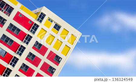 Vibrant modern building facade with red and yellow geometric patterns at a diagonal angle against a clear blue sky, featuring copy space for creative architectural design and urban concept. Vibrant modern building facade with red and yellow geometric patterns at a diagonal angle against a clear blue sky, featuring copy space for creative architectural design and urban concept. 136124549