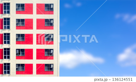 Modern residential building facade with red and white geometric patterns against a clear blue sky, featuring copy space for minimal architectural exterior design and urban living concept. Modern residential building facade with red and white geometric patterns against a clear blue sky, featuring copy space for minimal architectural exterior design and urban living concept. 136124551