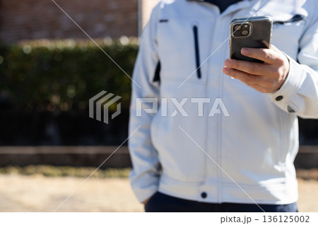 作業着を着た男性がスマホをいじりながらポケットに手を入れている 作業着を着た男性がスマホをいじりながらポケットに手を入れている 136125002