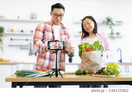 An Asian couple records a cooking video in their bright, modern kitchen, showcasing fresh ingredients 136125802