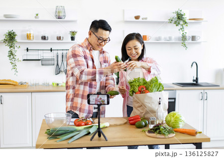 An Asian couple prepares fresh vegetables in their kitchen while filming a cooking tutorial for their online audience An Asian couple prepares fresh vegetables in their kitchen while filming a cooking tutorial for their online audience 136125827