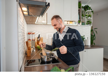 Happy man cooking meal adding fresh herbs in kitchen 136127150