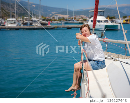 Mature woman relaxes on private yacht during sunny day in Finike Marina. Turkish coastline and luxury boats surround sailing vessel in Mediterranean port during summer vacation. Mature woman relaxes on private yacht during sunny day in Finike Marina. Turkish coastline and luxury boats surround sailing vessel in Mediterranean port during summer vacation. 136127448