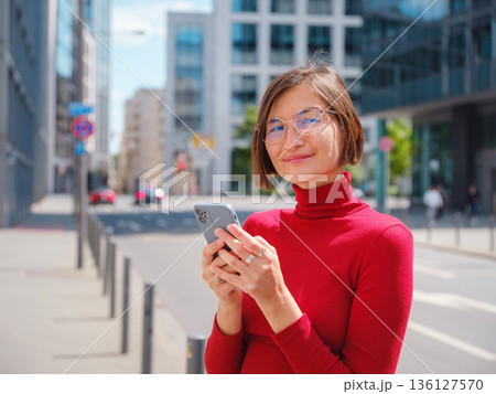 Beautiful woman in walking in Frankfurt on spring day and holding mobile phone. Urban lifestyle concept. Check social networks, send sms or book hotel, exuding confidence and professionalism Beautiful woman in walking in Frankfurt on spring day and holding mobile phone. Urban lifestyle concept. Check social networks, send sms or book hotel, exuding confidence and professionalism 136127570