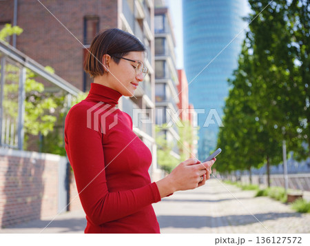 Beautiful woman in walking in Frankfurt on spring day and holding mobile phone. Urban lifestyle concept. Check social networks, send sms or book hotel, exuding confidence and professionalism 136127572