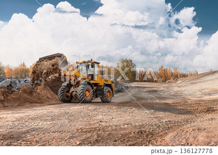 A loader is lifting and moving dirt at a construction site. The ground is uneven with rocks and material. Trees are visible in the background under a cloudy sky A loader is lifting and moving dirt at a construction site. The ground is uneven with rocks and material. Trees are visible in the background under a cloudy sky 136127778