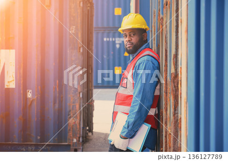 African American logistic workers wear yellow helmets and reflection shirts, hold document paper and look to check inventory or job details at the shipping container yard. Transportation import and African American logistic workers wear yellow helmets and reflection shirts, hold document paper and look to check inventory or job details at the shipping container yard. Transportation import and 136127789
