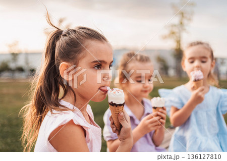 Three little girls enjoying ice cream cones in a park during sunset Three little girls enjoying ice cream cones in a park during sunset 136127810