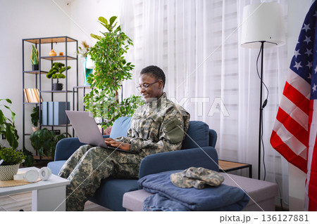 American female soldier working from home using laptop, with United States flag in background 136127881