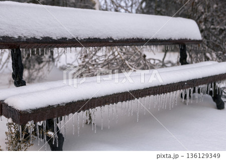an icy, snow-covered park empty bench with icicles hanging down 136129349