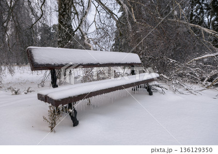 an icy, snow-covered park empty bench with icicles hanging down 136129350