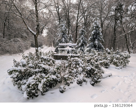 In a snowy winter park, a sculpture shaped like a bench with clothes stands beneath falling snow. In a snowy winter park, a sculpture shaped like a bench with clothes stands beneath falling snow. 136129364