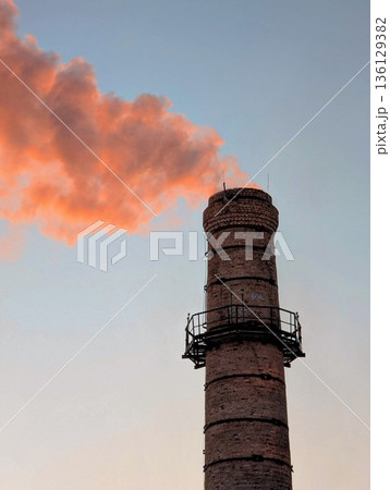 a heating plant chimney with a column of smoke pouring out, illuminated by the orange setting sun a heating plant chimney with a column of smoke pouring out, illuminated by the orange setting sun 136129382