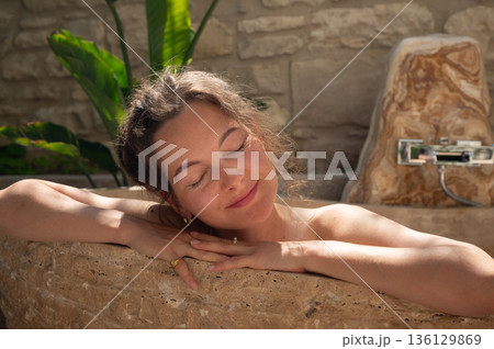 Serene woman relaxing in luxurious travertine bathtub enjoying spa treatment 136129869