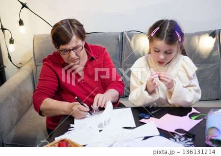 Grandmother and granddaughter create paper crafts together, enjoying a cozy moment of shared creativity. 136131214