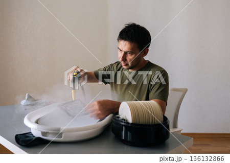Serious focused young man using steam cleaner to clean air humidifier filter in basin at home, ensuring regular maintenance for maintaining healthy indoor air quality. Concept of domestic work. Serious focused young man using steam cleaner to clean air humidifier filter in basin at home, ensuring regular maintenance for maintaining healthy indoor air quality. Concept of domestic work. 136132866