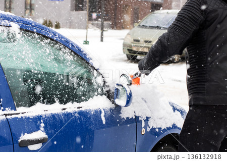 Individual in gloves brushing snow from vehicle on frosty residential street under gray sky 136132918