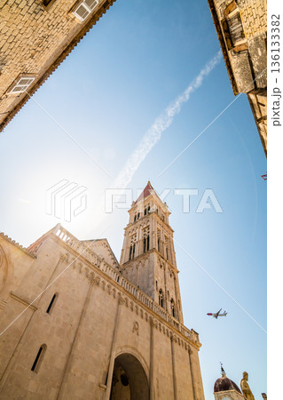 View of cathedral of St Lawrence, Trogir - Croatia. Down to up look to beautiful ancient church tower. The airplane is landing to Split airport. Aircraft is visible near the tower. View of cathedral of St Lawrence, Trogir - Croatia. Down to up look to beautiful ancient church tower. The airplane is landing to Split airport. Aircraft is visible near the tower. 136133382