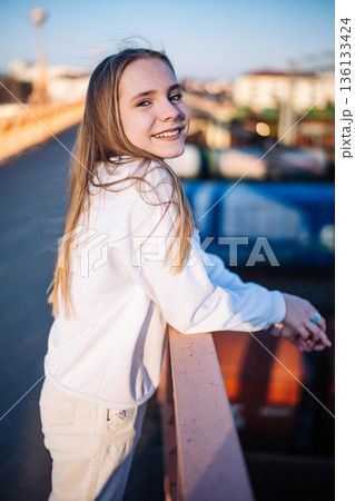 Happy teenage girl smiling, enjoying urban view on bridge at golden hour 136133424