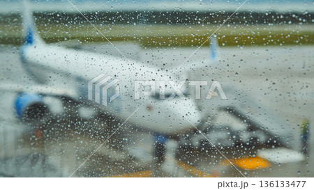 Raindrops blanketing airport window glass, blurring view of parked airplane, conveying travel anticipation and momentary stillness 136133477