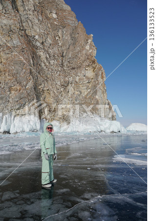 Woman in warm winter clothing stands on clear ice near massive frozen cliffs of Lake Baikal under bright blue sky Woman in warm winter clothing stands on clear ice near massive frozen cliffs of Lake Baikal under bright blue sky 136133523