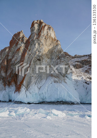 Massive rocky cliff rises from the frozen surface of Lake Baikal, covered with thick ice formations and snow under a clear blue winter sky 136133530