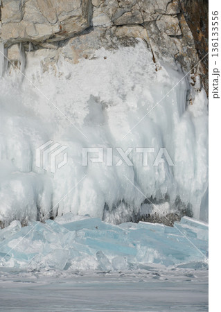 Sharp turquoise ice shards lie on the frozen surface of Lake Baikal with massive ice-covered cliffs in the background under clear winter light 136133556