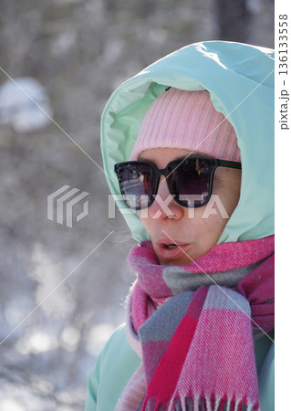 Close-up portrait of a woman in winter clothing with hood, scarf, and sunglasses, visible breath in cold air and soft snowy background Close-up portrait of a woman in winter clothing with hood, scarf, and sunglasses, visible breath in cold air and soft snowy background 136133558