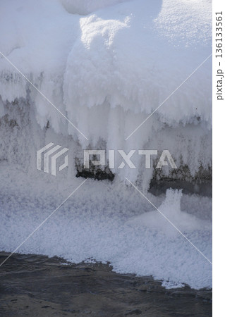 Frozen stream edge with thick snow layer and hanging ice formations above clear flowing water, soft winter light and calm mood 136133561