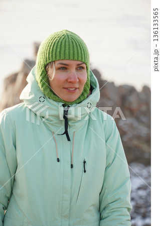 Smiling woman in a green knit hat and warm winter jacket stands outdoors near Lake Baikal in soft cold light 136133565