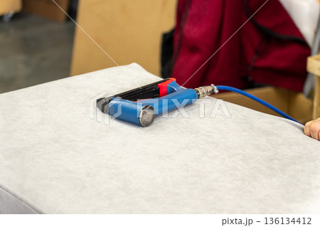 A worker uses a pneumatic stapler to attach white fabric to a furniture piece 136134412