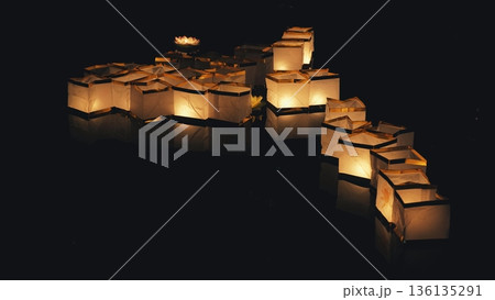 People sending glowing paper lantern boats onto a dark reflective river during a night festival. 136135291