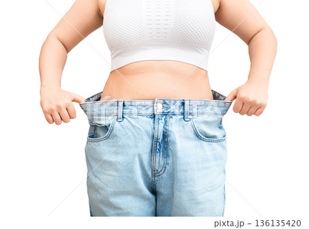 Slim woman showing weight loss success in oversized jeans. Fit female pulling loose pants away from waist isolated on white background 136135420