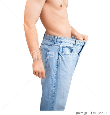 Slim man in oversized jeans showing successful weight loss results. Fit male pulling loose pants away from stomach isolated on white background 136135422