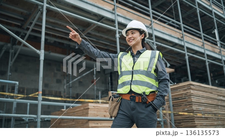 女性がヘルメットと安全ベストで足場前を指し示す日本の現場 136135573
