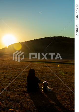 A woman sits in a field with her dog, watching the sun set A woman sits in a field with her dog, watching the sun set 136135778